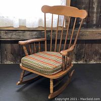 Side angled view of the wooden Roxton-style rocking chair showing the curved armrest, turned front leg, and patterned seat cushion.