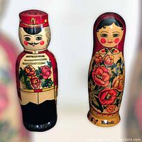 Front view of two Matreshka dolls, one female with floral design and one male in red uniform holding rodnik vodka bottle.