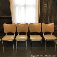 Front view of four vintage vinyl and chrome kitchen chairs, in tan color, placed in a row in front of a window with white lace curtains.