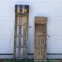 Two wooden vintage toboggans placed upright against a white garage door, showing front sides with straps and rope visible