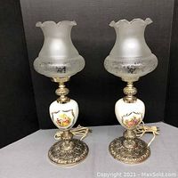 Front view of pair of decorative table lamps showing frosted scalloped glass shades, floral etched design on glass, ornate metal bases with tarnishing, and painted porcelain midsections with floral motifs