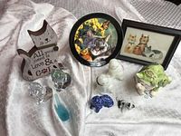 Photo showing two crystal cats, multiple cat figurines, wooden sign, clay trays, cat painting, and tabby cat plate with holder all arranged on white cloth