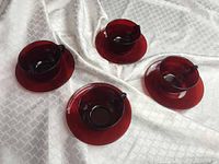 Four ruby red glass teacups, each paired with a matching saucer, sitting on a white textured fabric background. Cups have handles and show deep transparent ruby red color. One cup is noticeably thicker glass than the others.
