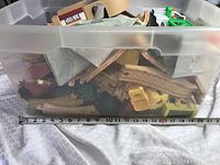 Plastic bin full of assorted wooden train tracks, trains, and accessories, mostly light-colored wood with a folded play mat.