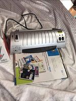 Silver Scotch thermal laminator powered on and resting on cloth surface with packaging boxes and laminating pouch packages below.