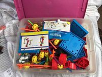 Top view of plastic bin filled with colorful Tinker Toys parts, including wheels, rods, connectors, flat plates, and instruction booklets.