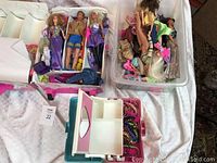 Multiple Barbie and Ken dolls arranged in boxes, with clothes showing different styles and patterns, alongside a pink travel case and a jewelry box with beads.