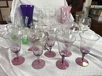 Various glassware items including several pink sherbet goblets with twisted spiral design, clear wine glasses and tumblers, slender green vases, a tall purple vase, and a pink vase arranged on a white cloth.