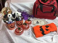 Full lot arranged on white fabric: glass pumpkins, ceramic buckets, stuffed Snoopy toys, string of purple cat lights, canvas pumpkin bag, Hallmark plate, red Delsey soccer bag, Halloween cloth with jack-o'-lantern face, and a Christmas musical globe.