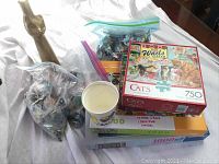 Photo showing two gallon-sized bags of Red Rose figures, four puzzle boxes stacked, a Mardi Gras cup, and a gold cat wine bottle statue.