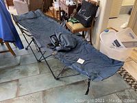 Foldable camp cot fully extended on floor showing dark fabric stretched on metal frame, indoors on tiled floor among furniture.