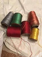 Six wooden spools of silk thread in gray, green, copper, red, burgundy, and yellow arranged on a fabric surface.