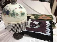 Boudoir lamp with floral reverse painted glass shade, fringe intact, two Native American woven textiles behind it.