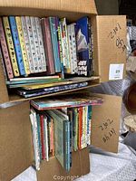 Two cardboard boxes filled with vintage children's books and story collections from the 1950s and 1960s, showing various colorful spined books stacked upright and layered.