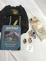 Photo showing full lot including black Harry Potter backpack with Hogwarts emblem patch, Hogwarts Journal book with stickers, drawstring bag for puzzle, some glass stones, worry dolls in a small basket, and magic trick box.