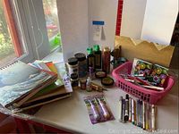 Overview of artist supplies including sketch pads, jars of pigment, glitter containers, pens, and a pink basket holding markers and pens on a white table near window.