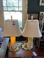Pair of matching Aladdin lamps on wooden table, showing lime green pudding glass bases, white fabric square-shaped shades, and original finials.