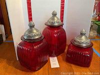 Set of three red ribbed Artland glass canisters with silver flower-embossed metal lids against a white background and measuring scale, showing all three canisters together.