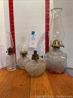 Four vintage Wexford by Anchor Hocking oil lamps shown side by side on a wooden surface with a measuring backdrop. Three have glass chimneys, one does not. Bases have diamond-cut pattern.