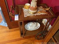 Front angled view of wooden tea cart with glass top, decorative scroll handle, and porcelain dish below.