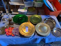 Overall view of assortment showing multiple green and silver plate chargers, two silver candelabra holders, colorful ceramic flower-shaped dishes, and metal pitcher.