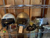 Two motorcycle helmets (Bell II open-face and Nolan full-face) along with various motorcycle accessories including goggles, sunglasses, face shields, and headphones on metal shelving.