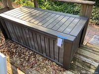 Full view of brown plastic outdoor deck box with hinged lid and vertical panel design, placed on deck with dry leaves around.