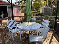 White round metal patio table with green umbrella and umbrella stand on a wooden deck with four matching white metal chairs showing paint wear and dirt.