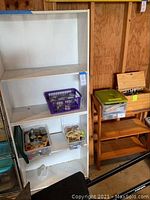 White tall wooden bookcase with three shelves and craft supply containers inside. Visible dust and wear from garage use.