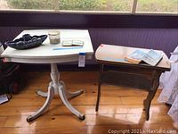 Photo of two tables side by side on wooden floor in enclosed porch, white pedestal table with foldable top and smaller brown wooden table with items on top
