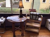 Full view of round wooden table with lamp on top, small wooden bookcase next to it with decorative box on top