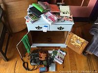 Photo of the small vintage cabinet with two front drawers, worn blue paint with chipped areas and missing knob, and items arranged around it including purses and a dish towel