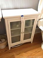 White wooden cabinet with frosted glass front doors, closed, standing on wooden floor near a wall.