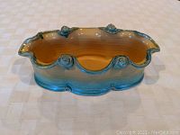 Side view of an oval hand thrown pottery bowl or planter showing scalloped rim and applied rose buds with blue exterior glaze and yellow interior.