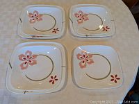 Four plates, two larger and two smaller, showing the pink floral design on white square plates with green curved lines, arranged on a beige checkered tablecloth.