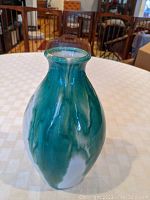 Front view of the green drip glaze art pottery vase showing its bulbous shape and flared rim on a table with a checkered tablecloth and background furniture.