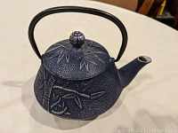 Side view of dark blue cast iron teapot with bamboo leaf and stalk design on body and lid, curved handle and spout.