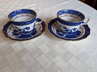 Two Royal Doulton Blue Willow teacups and matching saucers on a light-colored tablecloth from a top-side perspective.