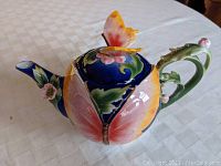 Side view of colorful butterfly ceramic teapot on white tablecloth showing butterfly wing design and flower accents on spout and handle.