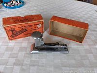 Pilot 402 stapler displayed next to its original orange box on a white checkered tablecloth, showing overall appearance and box design.