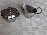 Photo shows a polished stainless steel creamer with curved handle and sugar bowl with square lid, both on a checkered fabric surface.
