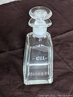 Front view of the clear glass oil and vinegar bottle with etched markings and glass stopper, displayed on dark fabric.