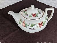 Angled side view of white ceramic teapot showing curved spout and handle with grapevine and berry decoration.