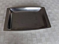 Top view of rectangular stainless steel serving platter placed on a table with slight reflections, showing flat broad rim and inner shallow tray surface with minor marks.