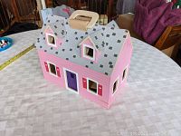 Closed view of the pink foldable wooden dollhouse with gray roof and handle on a table.