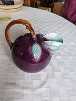 Plum-shaped purple ceramic pitcher with brown handle and green leaves on spout, photographed on white checkered tablecloth.