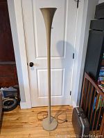 Full view of the beige mid-century modern torchiere floor lamp standing upright by a white door on wood flooring. Shows the slender stem and round base.