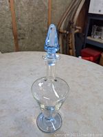 Full view of decanter showing clear body, sapphire blue foot and stopper on a table with other objects in background.