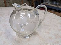 Side view of clear glass pitcher showing ball form body and etched apple motifs around the glass surface. Fluted handle attached on side.