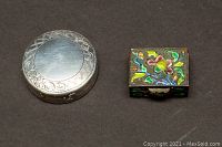 Closed view of two pill boxes: one round sterling silver with engraved designs, one rectangular with multi-colored enamel floral overlay.